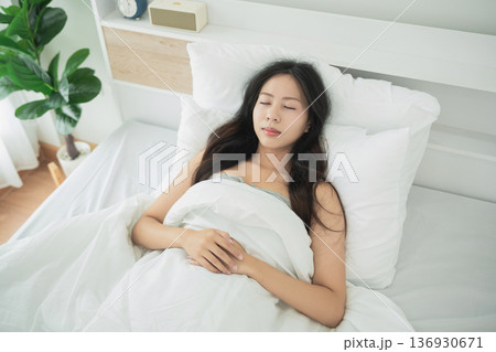 Peaceful young woman resting in bed, eyes closed, enjoying a moment of tranquility and relaxation under soft white blankets in cozy bedroom setting 136930671