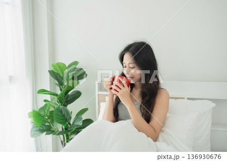 Young woman enjoying a warm beverage in a bright, cozy bedroom with white bedding and green plant in natural light for relaxation and comfort 136930676