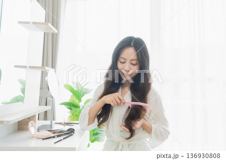 Young woman in light robe combing her long hair in a bright and airy room with natural light and greenery decorating the space Young woman in light robe combing her long hair in a bright and airy room with natural light and greenery decorating the space 136930808