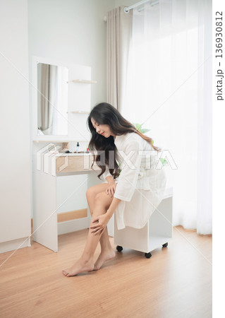 Young woman sitting on stool in bright room touching knee with pained expression during self-care routine in cozy, modern interior setting Young woman sitting on stool in bright room touching knee with pained expression during self-care routine in cozy, modern interior setting 136930812