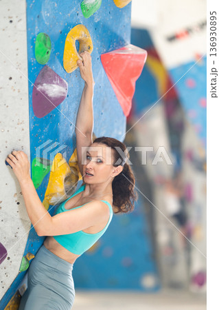 Adult woman is mastering climbing on training wall in gym, side view. Young woman holds on tightly to ledges and strives for top of bouldering route 136930895