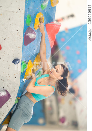 Adult woman is mastering climbing on training wall in gym, side view. Young woman holds on tightly to ledges and strives for top of bouldering route 136930915