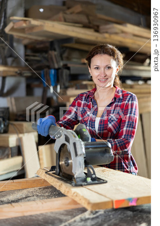 Female employee of workshop cuts wooden board into narrow parts using circular saw Female employee of workshop cuts wooden board into narrow parts using circular saw 136930967