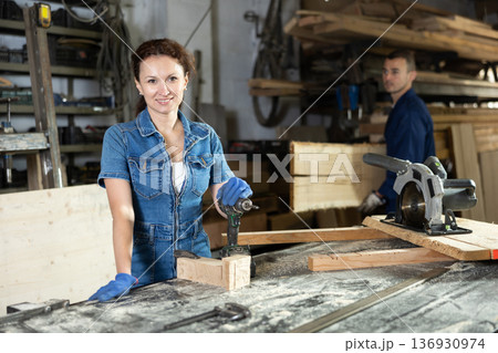 Female employee of carpentry workshop uses drill to make holes in wooden billet Female employee of carpentry workshop uses drill to make holes in wooden billet 136930974