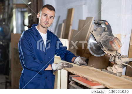 Male employee of workshop cuts wooden board into narrow parts using circular saw 136931029