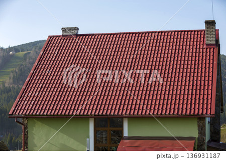 Detail of new modern house cottage corner with stucco walls decorated with natural stones, red shingled roof and rain gutter pipe system on blurred spruce forest copy space background. 136931187