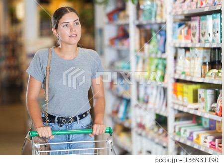 Young woman pushing shopping cart along grocery shelves in store Young woman pushing shopping cart along grocery shelves in store 136931190