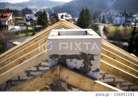 Close-up detail of roof frame of rough wooden lumber beams and chimney made of foam insulation blocks on blurred green background. Building, roofing, construction and renovation concept. Close-up detail of roof frame of rough wooden lumber beams and chimney made of foam insulation blocks on blurred green background. Building, roofing, construction and renovation concept. 136931191