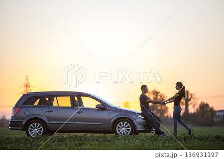 Young couple, slim attractive woman with long ponytail and handsome man standing at silver car in green field on clear sky at sunset or sunrise copy space background. Young couple, slim attractive woman with long ponytail and handsome man standing at silver car in green field on clear sky at sunset or sunrise copy space background. 136931197