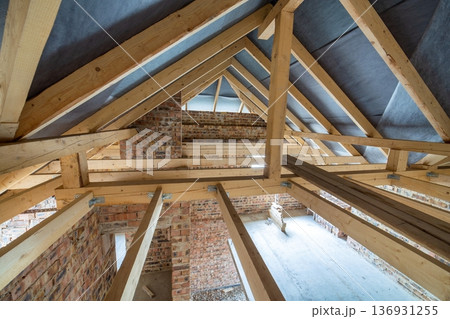 Attic space of a building under construction with wooden beams of a roof structure and brick walls. Real estate development concept. Attic space of a building under construction with wooden beams of a roof structure and brick walls. Real estate development concept. 136931255