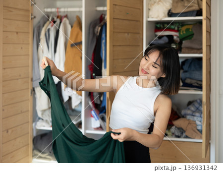 Happy young Asian girl trying to choose a dress standing by wardrobe Happy young Asian girl trying to choose a dress standing by wardrobe 136931342