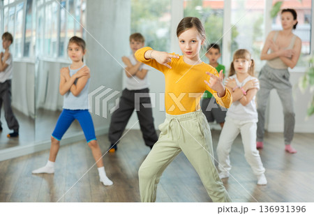 Positive juvenile girl engaged in breakdancing in training room with children's group 136931396