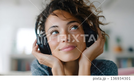 A thoughtful young woman with curly hair and freckles listens intently with black over-ear headphones, gazing upward while enjoying music or a peaceful podcast. AI Generated 136934301