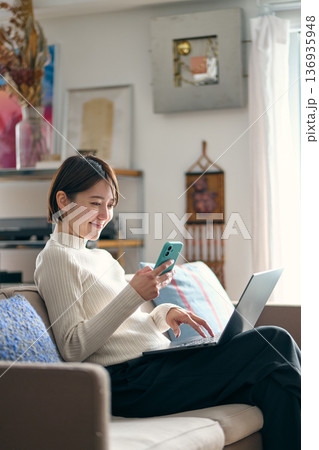 パソコンを操作しながらスマートフォンを手に持つ女性 136935948