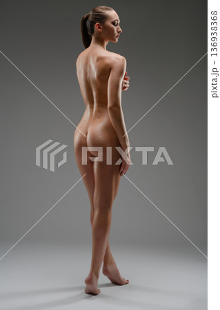 Elegant full-length nude woman posing from behind in studio. Athletic female body with graceful posture on a neutral gray background. Elegant full-length nude woman posing from behind in studio. Athletic female body with graceful posture on a neutral gray background. 136938368
