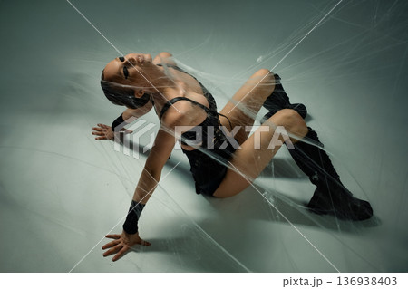 A woman in a black gothic outfit with dramatic makeup lies trapped and entangled in a large spiderweb on the floor. 136938403
