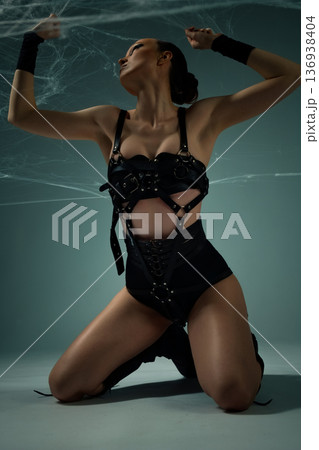 Powerful woman in a black leather harness kneeling under spiderwebs in a dramatic pose 136938404