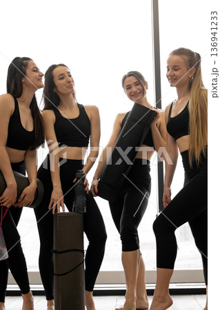 Four young women in athletic wear are holding yoga mats and water bottles, smiling and chatting after a workout session 136941233