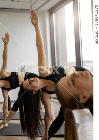 Three women in a yoga studio perform the triangle pose, stretching their bodies and focusing on their practice 136941270
