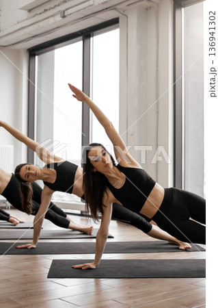 A group of women in black activewear perform a side plank exercise on yoga mats in a bright studio with large windows A group of women in black activewear perform a side plank exercise on yoga mats in a bright studio with large windows 136941302