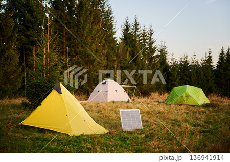 Solar panel in grassy field with tents in background highlights eco-friendly camping setup, surrounded by dense evergreen trees in evening. Integration of renewable energy in outdoor activities. Solar panel in grassy field with tents in background highlights eco-friendly camping setup, surrounded by dense evergreen trees in evening. Integration of renewable energy in outdoor activities. 136941914