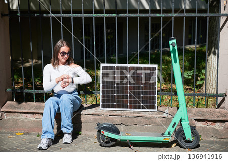 Woman checking her watch while charge electric scooter from portable solar panel, emphasizing integration of sustainable renewable energy. Concept of eco-friendly solutions in urban settings. 136941916