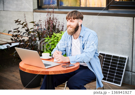 Bearded man works at round wooden table with laptop, notebook, and cup of coffee. Guy wears earbuds, surrounded by greenery and solar panel, highlighting modern, eco-friendly, productive workspace. Bearded man works at round wooden table with laptop, notebook, and cup of coffee. Guy wears earbuds, surrounded by greenery and solar panel, highlighting modern, eco-friendly, productive workspace. 136941927