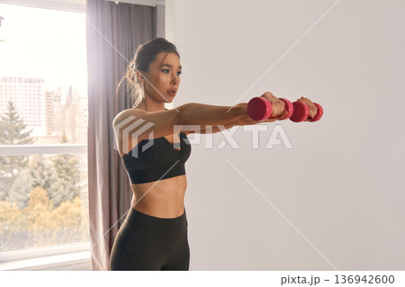 Woman performing strength training with pink dumbbells in a bright room while standing by large windows and enjoying the view 136942600