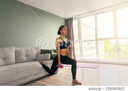 Fit woman doing lunges with dumbbell in modern living room during afternoon workout session Fit woman doing lunges with dumbbell in modern living room during afternoon workout session 136942601