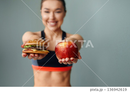 Woman holding an apple in one hand and a burger in the other while smiling against a simple background 136942619