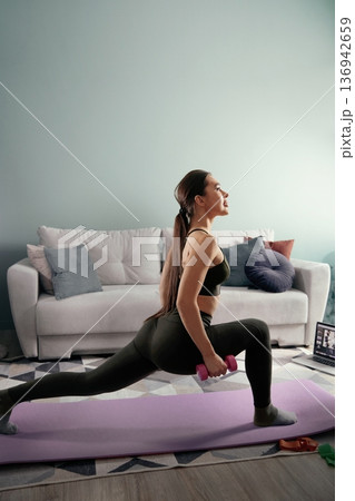 Woman exercises with weights in living room during daytime while following online workout routine Woman exercises with weights in living room during daytime while following online workout routine 136942659