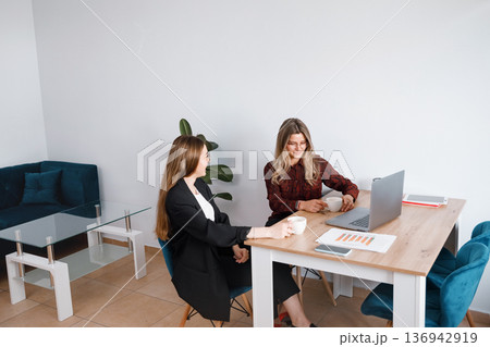 Women work together in office setting discussing ideas at a wooden table during daytime hours in a modern space Women work together in office setting discussing ideas at a wooden table during daytime hours in a modern space 136942919