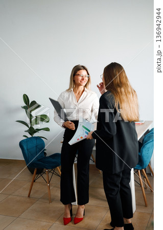Women discuss ideas in an office setting with charts and documents during a work session Women discuss ideas in an office setting with charts and documents during a work session 136942944