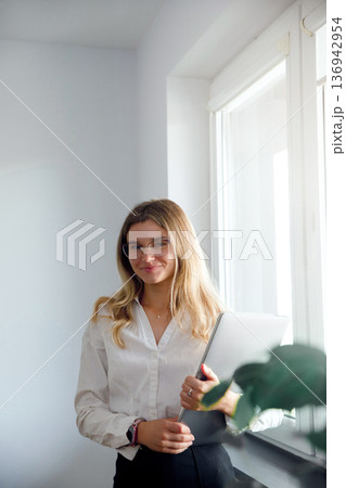Woman in white shirt stands in office by windows holding laptop and smiling during a workday Woman in white shirt stands in office by windows holding laptop and smiling during a workday 136942954