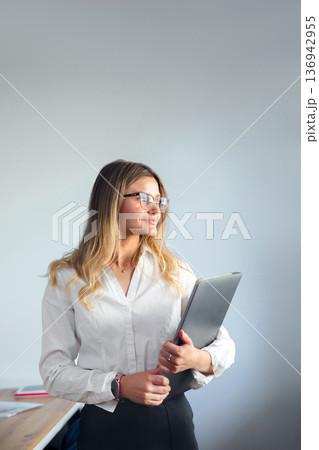 Businesswoman stands with a laptop while smiling in a bright office during the day Businesswoman stands with a laptop while smiling in a bright office during the day 136942955