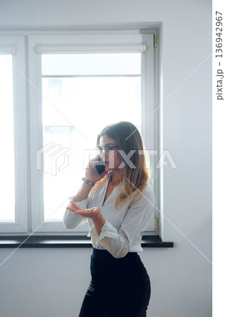 Businesswoman talking on phone while standing near window in office during work hours Businesswoman talking on phone while standing near window in office during work hours 136942967