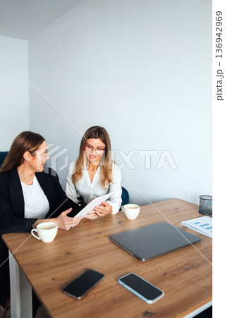 Women working together in an office discussing ideas during a meeting at a wooden table with laptops and coffee cups Women working together in an office discussing ideas during a meeting at a wooden table with laptops and coffee cups 136942969
