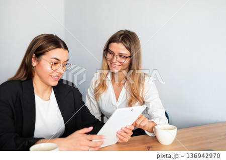 Women collaborate in modern office setting while discussing ideas and reviewing data on tablet during work hours Women collaborate in modern office setting while discussing ideas and reviewing data on tablet during work hours 136942970