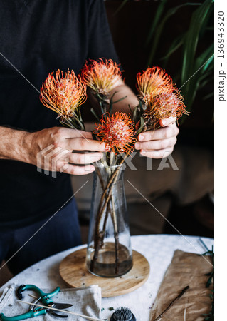 Person holds orange pincushion protea flower while arranging bouquet in glass vase on table. art therapy, tactile mindfulness, sensory wellness, therapeutic gardening Person holds orange pincushion protea flower while arranging bouquet in glass vase on table. art therapy, tactile mindfulness, sensory wellness, therapeutic gardening 136943320