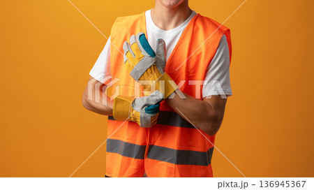Worker wears safety vest and gloves in studio with orange background for construction work Worker wears safety vest and gloves in studio with orange background for construction work 136945367