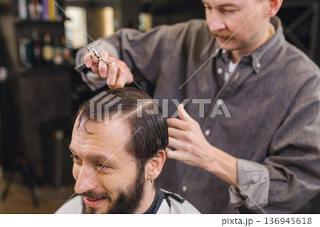 Barber cutting male client hair with scissors and comb. Close up of professional haircut process, precision technique, men grooming service in modern barbershop. 136945618