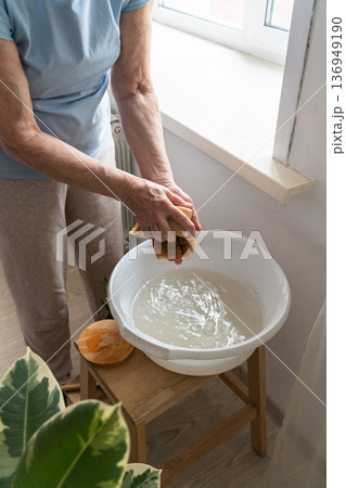 An elderly woman's hands holding bamboo cloth and natural soap for washing windows near houseplants, an authentic moment. An eco-friendly lifestyle, using natural, biodegradable cleaning products 136949190