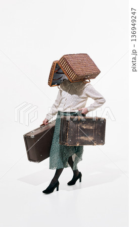 Woman in retro outfit carrying old suitcases with wicker basket covering head on white background 136949247