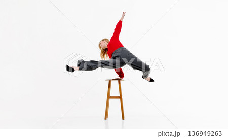 Female model in red sweater and gray trousers performing dramatic split leap over wooden stool in studio. Female model in red sweater and gray trousers performing dramatic split leap over wooden stool in studio. 136949263