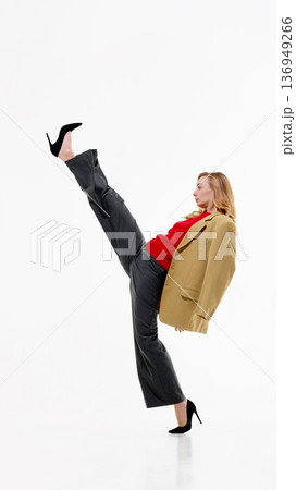 Woman in beige blazer, red sweater and gray trousers performing high kick on white background. Woman in beige blazer, red sweater and gray trousers performing high kick on white background. 136949266