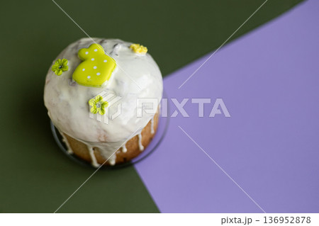 Flat lay of Easter cake topped with icing and pastel green bunny decoration on green-purple backdrop, modern holiday styling 136952878