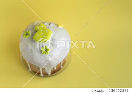 Easter kulich with glossy icing and light green bunny decoration on bright yellow background, cheerful seasonal styling 136952881