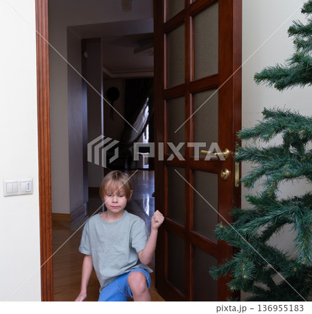 A young Caucasian boy with blonde hair is kneeling on the floor near a wooden door with glass panels. 136955183