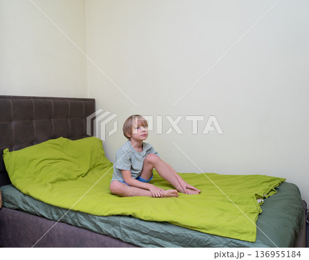 A young Caucasian boy with blonde hair sits barefoot on a bed with a bright green duvet. 136955184