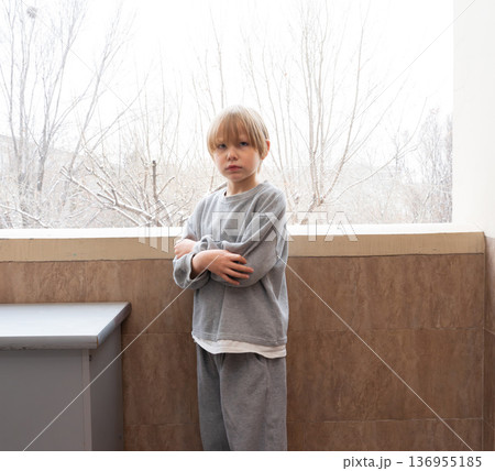 A young Caucasian boy with blonde hair stands on a balcony or porch with his arms crossed over his chest. 136955185